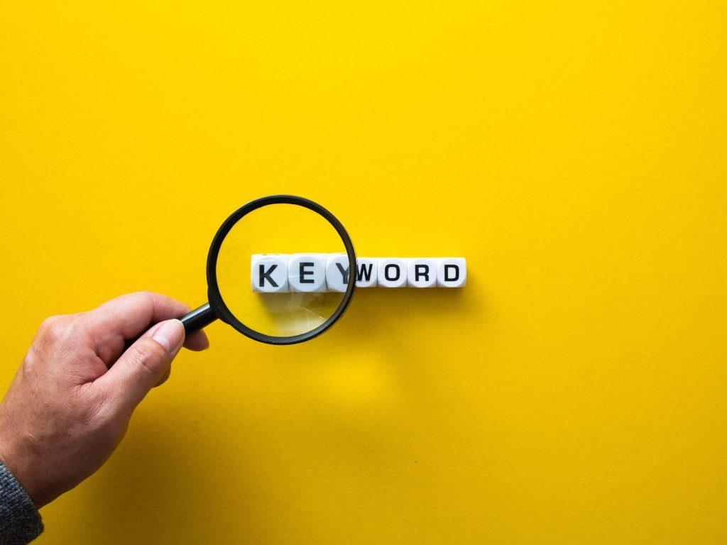 A hand holding a magnifying glass over letter blocks spelling KEYWORD on a bright yellow background