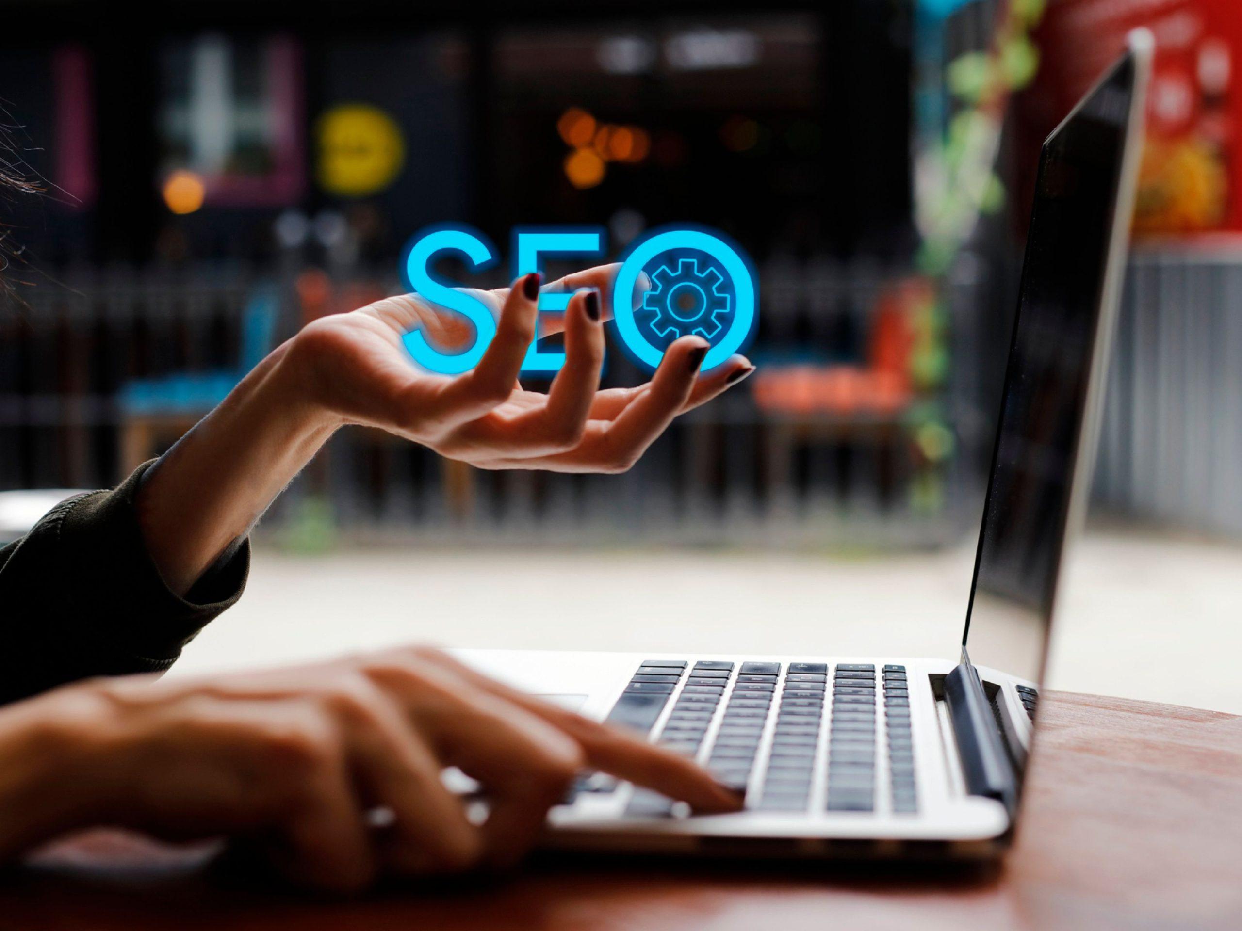 A hand hovering over a laptop keyboard, holding a glowing SEO icon with a gear