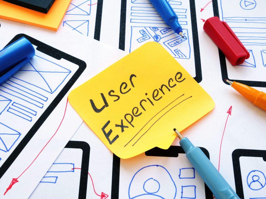 A sticky note labeled 'User Experience' on top of wireframe sketches and colorful markers